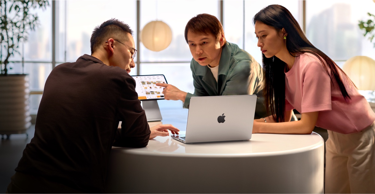三人聚在一张白色圆桌旁进行讨论。左侧的男子正在操作一台银色 MacBook Pro；中间的男子正用手指着旁边支架上一台 iPad 的屏幕，屏幕显示着收银系统的界面；右侧的女子正注视着 MacBook Pro 屏幕。