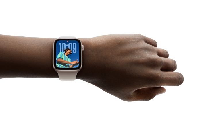 特写画面显示一只手腕戴着配有橡胶表带的玫瑰金色 Apple Watch，表盘展示全新 Liquid Glass 设计的时间数字。
