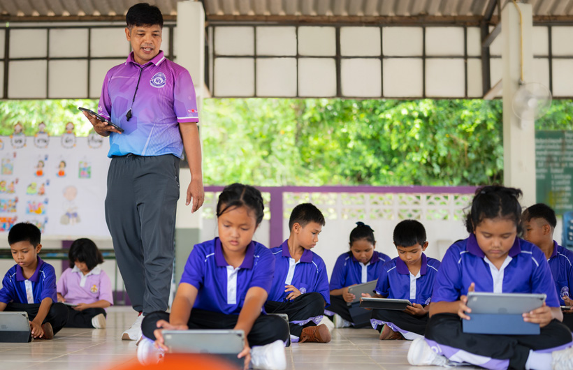 户外课堂上，学生们席地使用 iPad，一名体育老师在他们中间走动。
