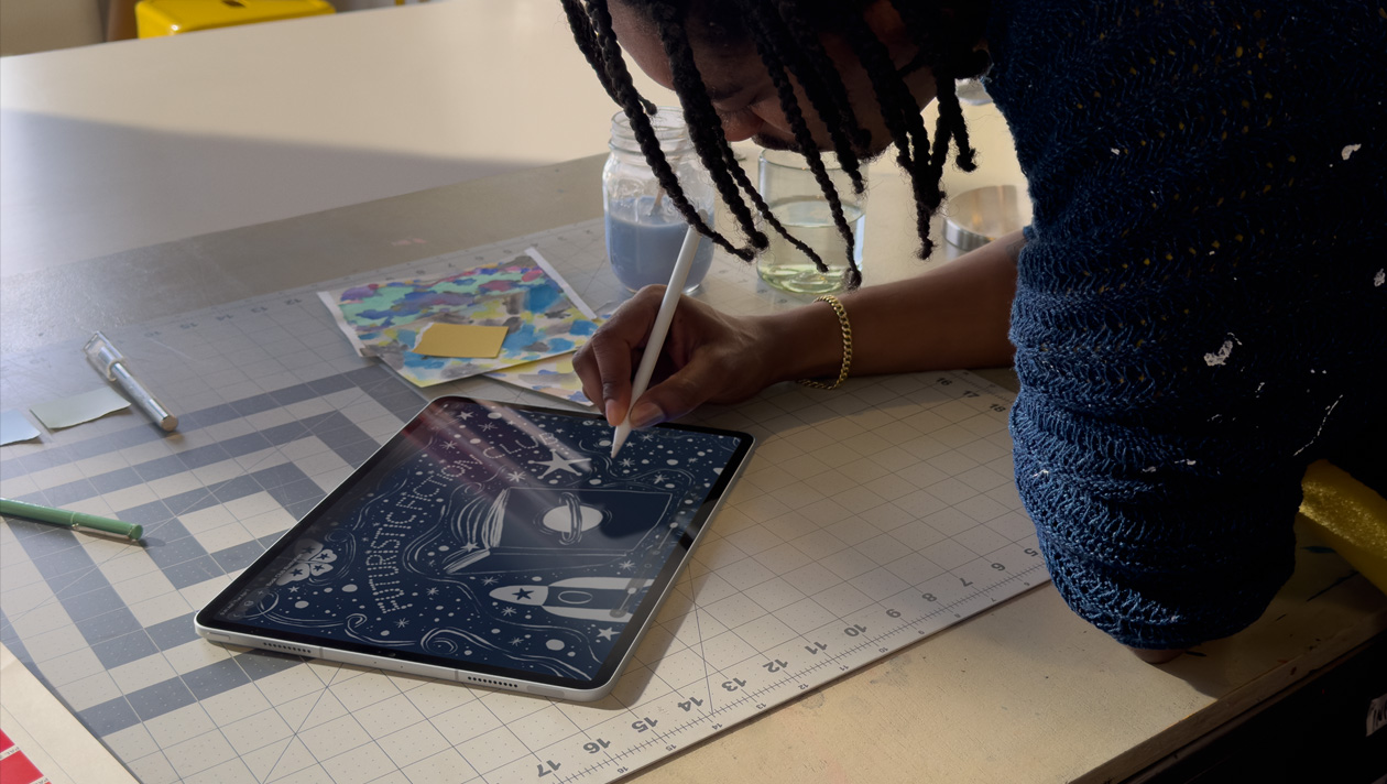 身穿蓝色毛衣、梳着辫发的人用 Apple Pencil 在 iPad Pro 上绘画，屏幕显示一幅夜空插画，其中包括封面有星球图案的书、宇宙飞船、蝴蝶，中间有文字“Futuristic Fiction Club”。